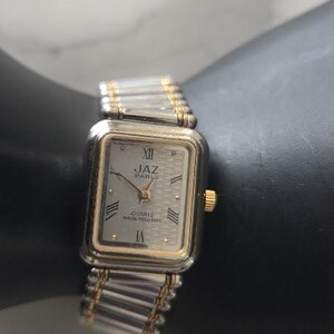 May include: A rectangular Jaz Paris quartz watch with a silver and gold-tone metal expansion band. The watch face is silver with a textured pattern, black Roman numeral hour markers, gold-tone hands, and the text Jaz Paris Quartz Water Resistant.