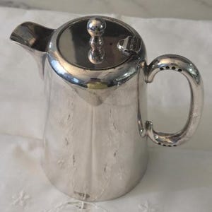 May include: A polished silver-toned metal creamer or small pitcher featuring a smooth, tapered body, a curved handle with decorative circular indentations, a short spout, and a circular lid topped with a small, rounded spherical finial.