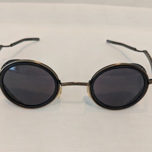 May include: A pair of round, black framed sunglasses with dark lenses. The frames are made of metal and have a steampunk aesthetic.