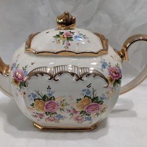May include: A white teapot with a gold trim and floral pattern. The teapot has a lid with a gold knob and a handle on the side.