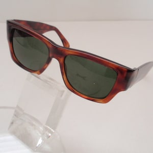May include: Tortoise shell sunglasses with green lenses. The sunglasses are on a clear display stand. The word "remarkable" is printed on the right lens.