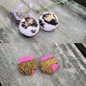 May include: Two pairs of round fabric earrings. One pair features a white background with black and gold abstract designs. The other pair has a pink top with a leopard print design. The earrings are displayed on a weathered wooden surface.