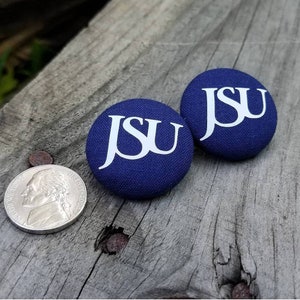 May include: Two navy blue fabric-covered buttons with the white letters "JSU" printed on them. A nickel coin is next to the buttons. The buttons are on a weathered wooden surface.