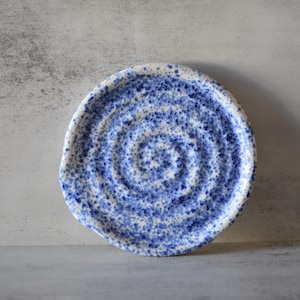 May include: A small, white ceramic dish with a blue and white speckled glaze. The dish has a slightly irregular shape and a swirling pattern.