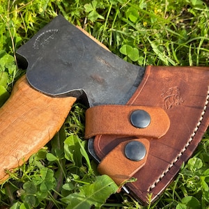 Könnte beinhalten: Eine braune Lederaxt-Scheide mit Druckknopfverschluss und einem Metallknopf. Die Scheide ist für eine kleine Axt mit einem Holzstiel konzipiert. Die Axt ist nicht im Bild enthalten.