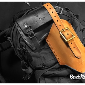 May include: A tan leather axe sheath with a brass buckle and a black leather strap. The sheath is attached to a black backpack with a zipper. The text "BushGear Leather Works" is visible on the sheath.