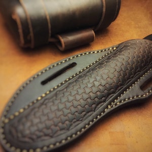Può includere: Un fodero per coltello in pelle marrone con un motivo di cuciture decorative e una chiusura a lembo.