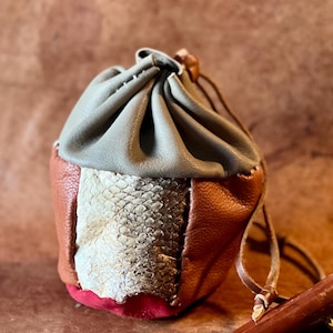Puede incluir: Una pequeña bolsa de cuero con cordón de ajuste, con la parte superior gris, la inferior marrón y un panel de piel de serpiente. La bolsa tiene un forro rojo y un cordón de cuero marrón.