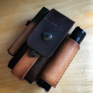 May include: A brown leather multi-tool pouch with a tan leather strap and a black flashlight. The pouch has the text "BushGear" and "Leather Works" embossed on it.