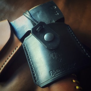 May include: Close-up of a black leather axe sheath with a brown leather strap and a metal button. The sheath has the text "Bush Craft" embossed on it.