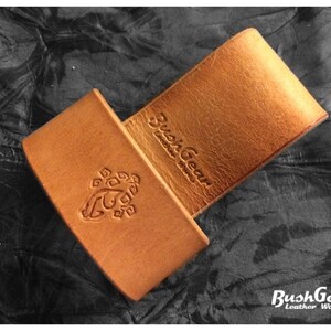 May include: A brown leather knife sheath with a BushGear Leather Works logo embossed on the front. The sheath has a belt loop on the back.