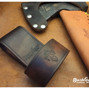 Puede incluir: Hacha y accesorios de cuero. La imagen muestra una cabeza de hacha marrón oscuro con una funda de cuero negra y dos fundas de cuero marrón. Las fundas están grabadas con el texto "BushGear Leather Works".