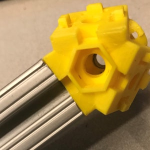 Op de afbeelding: Een heldergele, geometrische 3D-geprinte component bevestigd aan twee parallelle, zilverkleurige metalen staven. De component heeft een bolvorm met zeshoekige openingen en een getextureerd oppervlak.