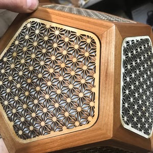 Op de afbeelding: Een houten geometrische doos met ingewikkelde stervormige uitsparingen. De doos heeft een lichtbruine afwerking met een contrasterende crèmekleurige achterkant achter de uitsparingen. Het ontwerp is symmetrisch en gedetailleerd.