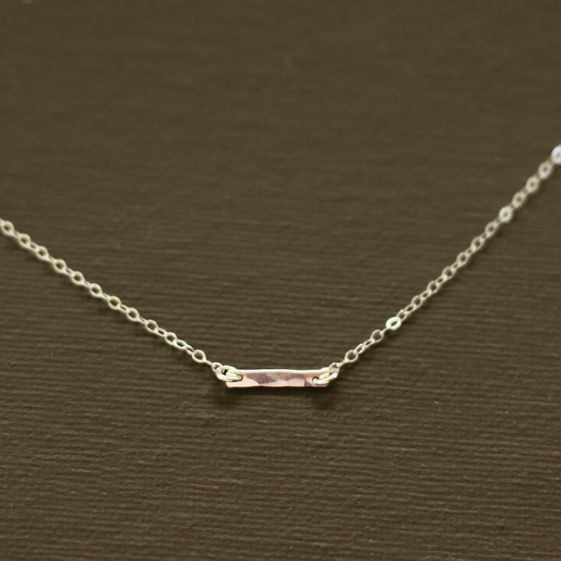 May include: A delicate silver necklace with a hammered bar pendant. The chain is a simple, linked style.