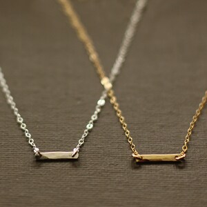 May include: Two delicate necklaces with thin chains and small, flat bar pendants. One necklace has a silver pendant and the other has a gold pendant.