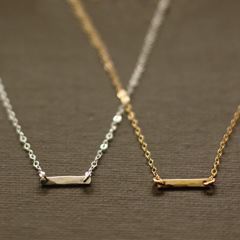 May include: Two delicate necklaces with thin chains and small, flat bar pendants. One necklace has a silver pendant and the other has a gold pendant.