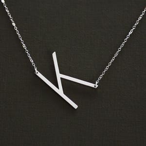 May include: A silver necklace with a minimalist letter K pendant. The pendant is made of a single piece of metal and has a simple, geometric design.