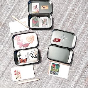 May include: Three rectangular metal tins with hinged lids, decorated with vintage-style postage stamps, floral motifs, and cross designs. Two small white cards with botanical illustrations and two light-colored pencils are arranged alongside the tins.