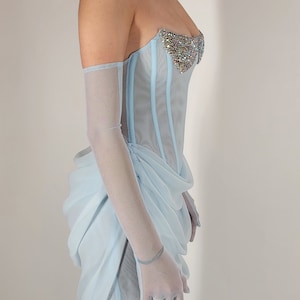 May include: A light blue strapless corset with a beaded embellishment on the bust. The corset is made of a sheer fabric and has a boned structure. The model is wearing the corset with a matching light blue skirt.