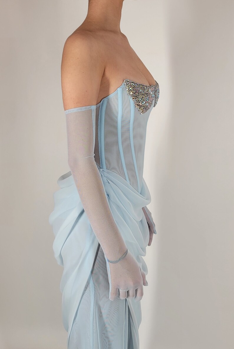 May include: A light blue strapless corset with a beaded embellishment on the bust. The corset is made of a sheer fabric and has a boned structure. The model is wearing the corset with a matching light blue skirt.