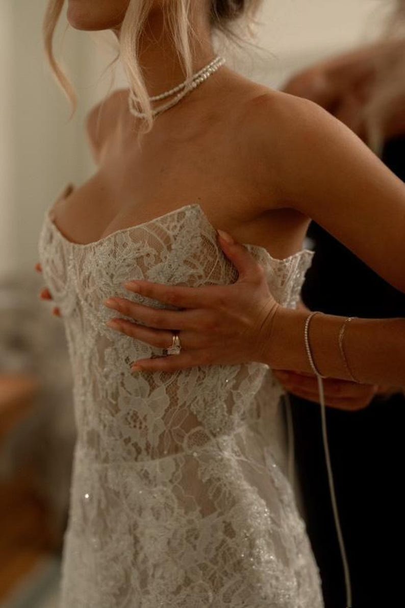 May include: A white lace wedding dress with a sweetheart neckline and a fitted bodice. The dress has a beaded embellishment and a flowing skirt.