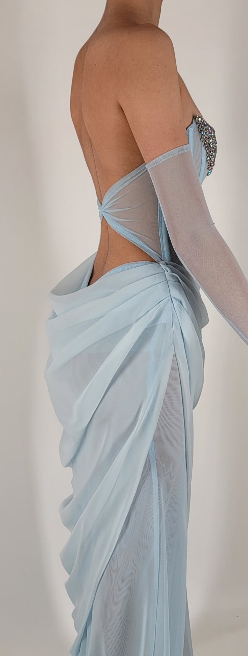 May include: A light blue, sheer, sleeveless gown with a low back and a gathered skirt. The gown has a fitted bodice with a sweetheart neckline and a beaded embellishment at the bust.