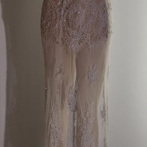 May include: A white lace gown with a fitted bodice and a flowing skirt. The gown has a lace-up back and is decorated with sequins. The gown is sheer and reveals the legs of the mannequin.