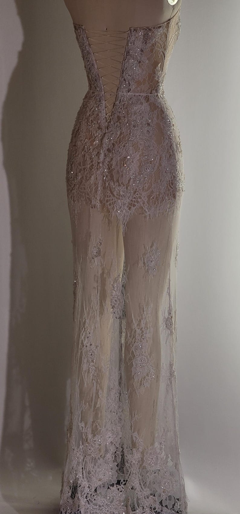 May include: A white lace gown with a fitted bodice and a flowing skirt. The gown has a lace-up back and is decorated with sequins. The gown is sheer and reveals the legs of the mannequin.