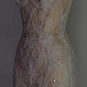 May include: A white lace dress with a fitted bodice and a flowing skirt. The dress is embellished with small, white beads and sequins. The dress is displayed on a mannequin against a gray background.