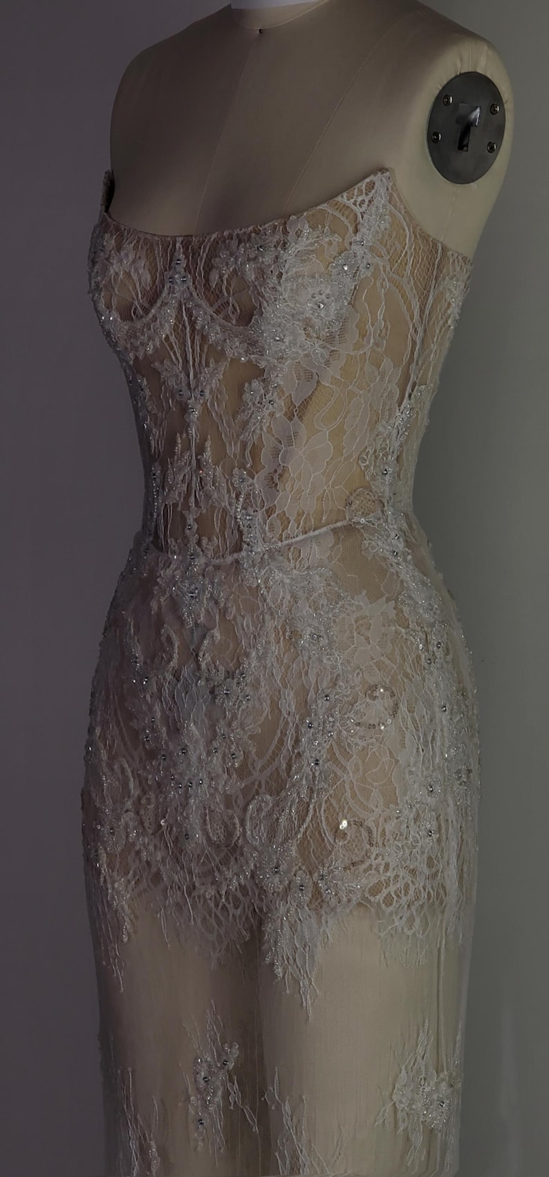 May include: A white lace dress with a fitted bodice and a flowing skirt. The dress is embellished with small, white beads and sequins. The dress is displayed on a mannequin against a gray background.