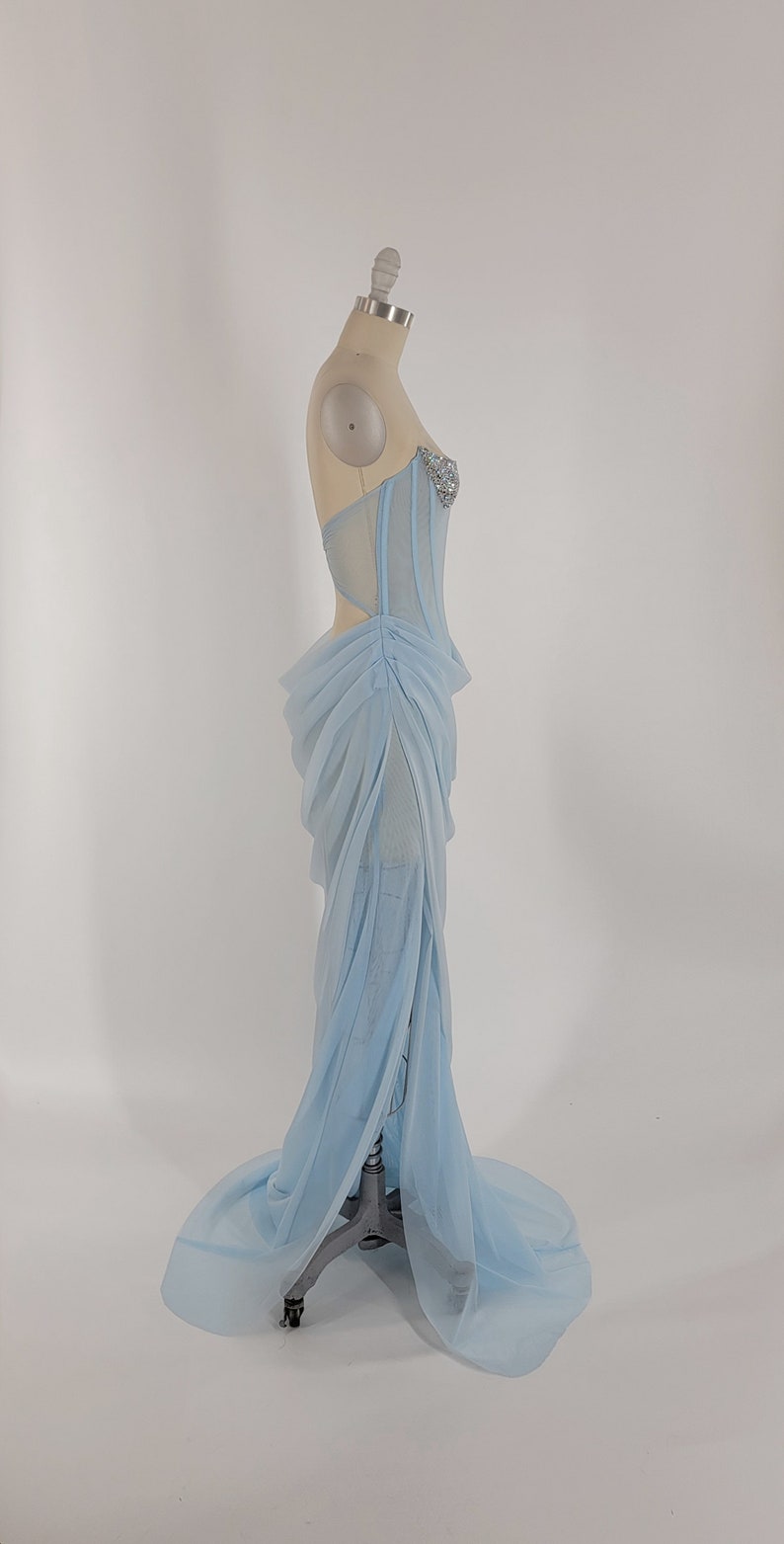 May include: A light blue, sheer, long, formal gown with a fitted bodice and a flowing skirt. The bodice has a sweetheart neckline and is adorned with rhinestones.