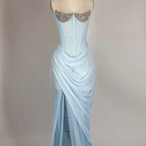 May include: A light blue strapless gown with a sheer bodice adorned with iridescent rhinestones. The skirt is made of flowing fabric with a draped design.