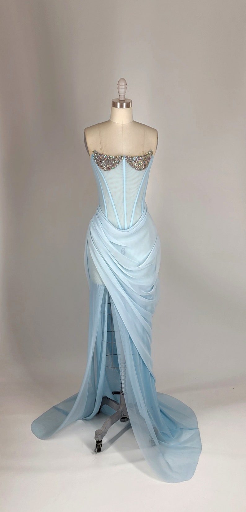 May include: A light blue strapless gown with a sheer bodice adorned with iridescent rhinestones. The skirt is made of flowing fabric with a draped design.