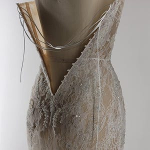 May include: A white lace wedding dress with a low back and a beaded design. The dress is on a white mannequin and the back of the dress is open with a zipper closure.
