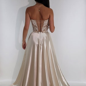 May include: A strapless, ivory satin gown with a lace bodice and a corset-style back. The gown has a full skirt and a train. The gown is on a white background.