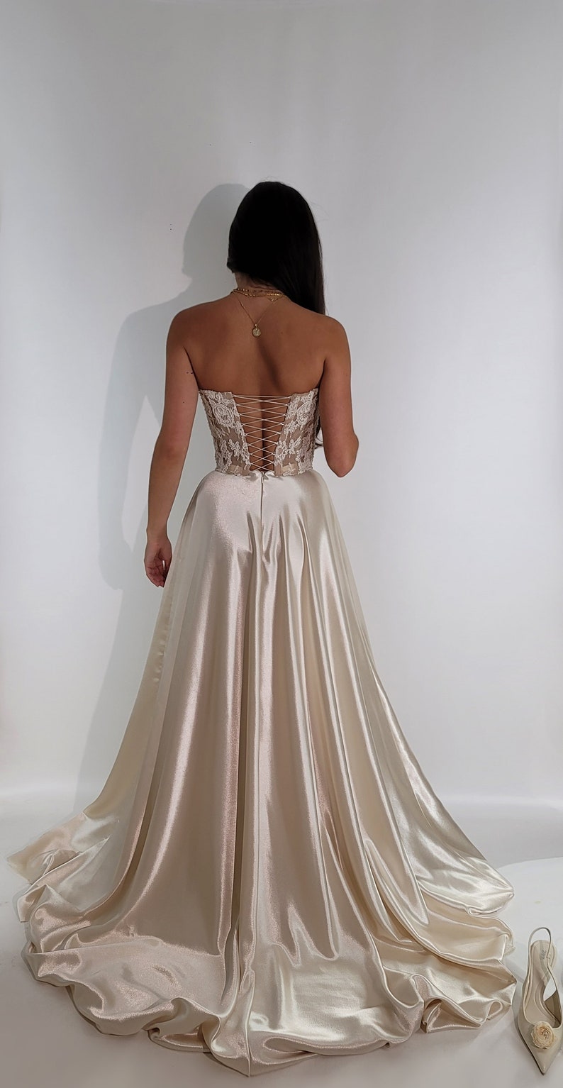 May include: A strapless, ivory satin gown with a lace bodice and a corset-style back. The gown has a full skirt and a train. The gown is on a white background.