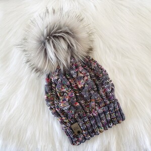 May include: A grey and multi-coloured knit beanie with a fluffy white pom pom. The beanie has a cable knit pattern and a small brown leather tag with the word "H&C" on it.