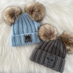 May include: Two crocheted beanies with faux fur pom poms. The beanie on the left is light blue with a black bear patch that says "Baby Bear". The beanie on the right is grey with a moose patch.