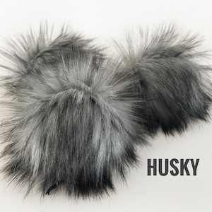 May include: Two fluffy gray and black faux fur pom poms. The larger pom pom is on the left and the smaller pom pom is on the right. The word "HUSKY" is printed in black below the pom poms.