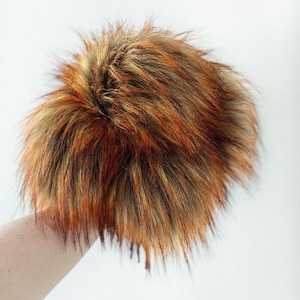 May include: A fluffy brown and orange faux fur pom pom. The pom pom is made of soft, plush fabric and has a long, shaggy texture.