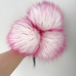May include: Two fluffy, pink and white faux fur pom poms against a white backdrop. The fur has a long pile with a vibrant pink hue at the tips, blending into a white base. The pom poms are a soft, textured accessory.