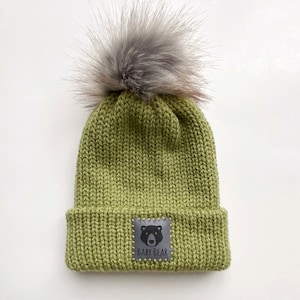 Peut inclure: Un bonnet en tricot vert avec un pompon en fausse fourrure sur le dessus. Le bonnet a un patch en cuir noir avec une silhouette d'ours et le texte "BABY BEAR" dessus.