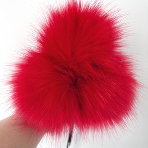 May include: Bright red fluffy pom-poms. The pom-poms are made of soft, textured material. The image shows two pom-poms, with a black string attached to them. The background is white.
