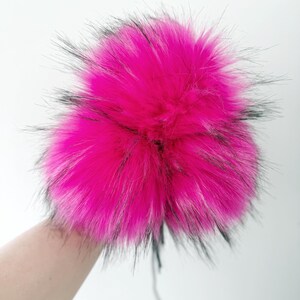 May include: A round, fluffy pom-pom crafted from vibrant hot pink faux fur with long, wispy black-tipped fibers extending from the center. The texture is dense and voluminous, creating a spherical shape with a bright, saturated color profile.