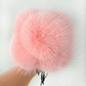 May include: A light pink fur hat with a fluffy texture. The hat is round and appears soft to the touch. Black ties are attached to the bottom. The hat is held by a person's arm.