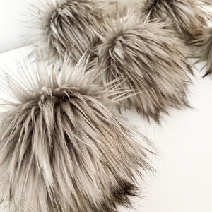 May include: A collection of four round, fluffy faux fur pom-poms in a variegated taupe and cream color palette. Each pom-pom features long, wispy synthetic fibers with darker brown tips, creating a textured, spherical appearance for craft projects.