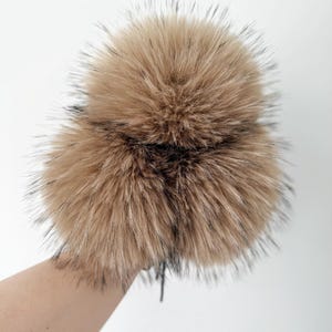 May include: A fluffy, round pom-pom accessory featuring long, soft fibres in a warm tan colour with dark brown or black tips. The texture is dense and voluminous, creating a spherical shape with a slightly spiky appearance at the edges.
