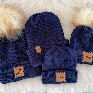 May include: Four navy blue knit beanies with faux fur pom-poms. Each beanie has a brown leatherette patch with text: "mama", "dada", and "mini". The hats are arranged on a white fluffy surface.