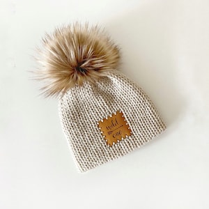 May include: A beige knitted beanie with a faux fur pom pom and a brown leather patch that says "wild one" with an arrow.